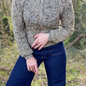 May include: A brown and beige crocheted jacket with a zip closure. The jacket has a textured pattern and is worn over a pair of dark blue jeans.