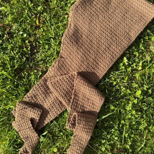 May include: A brown, crocheted scarf with a textured pattern. The scarf is long and rectangular, with a hood-like shape at one end and a pointed tip at the other. The scarf is lying on a bed of green grass.