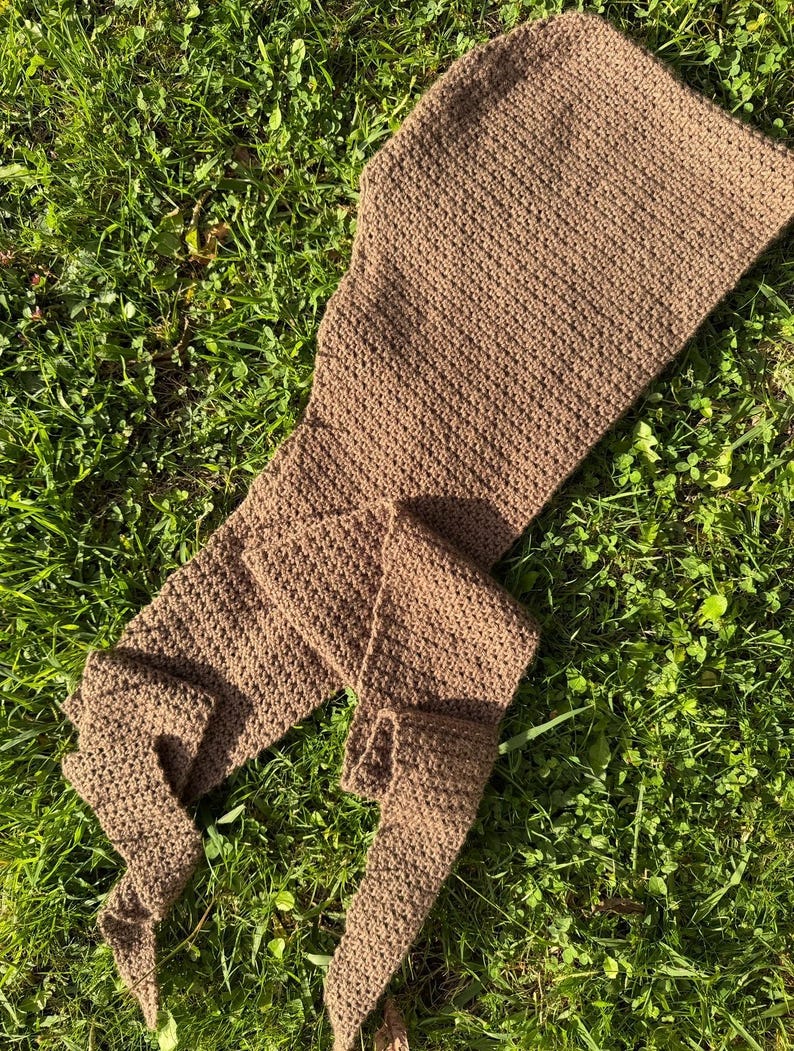 May include: A brown, crocheted scarf with a textured pattern. The scarf is long and rectangular, with a hood-like shape at one end and a pointed tip at the other. The scarf is lying on a bed of green grass.