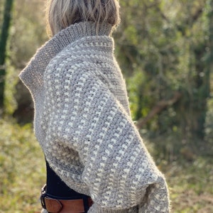 Peut inclure: Un pull en crochet gris et blanc avec un col roulé et des manches longues. Le pull est réalisé avec un point texturé et a une coupe ample.