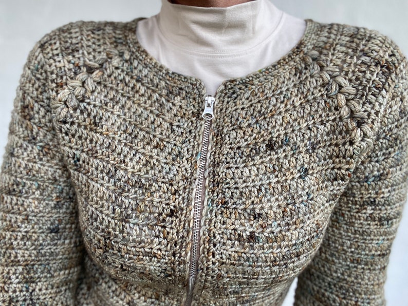 May include: A brown, green and white crocheted cardigan with a zip fastening. The cardigan has a textured stitch pattern and a round neckline.