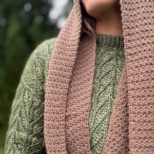 May include: A close-up of a person wearing a green sweater and a brown crocheted scarf and hood. The sweater has a diamond pattern. The scarf and hood are a solid color.