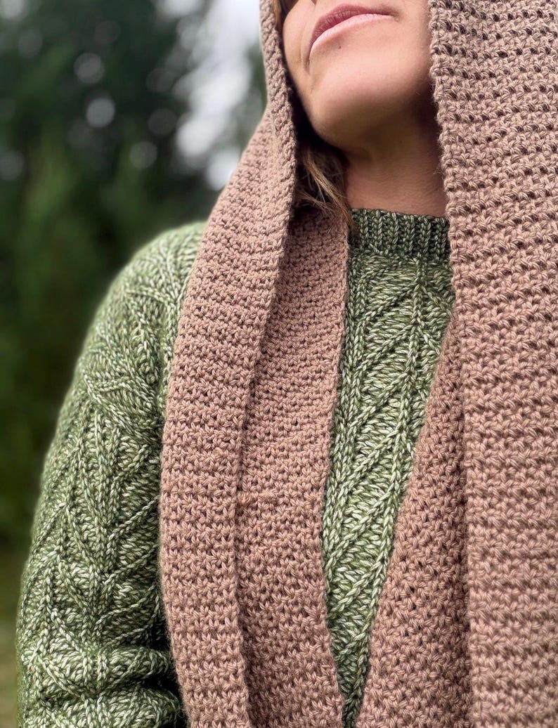 May include: A close-up of a person wearing a green sweater and a brown crocheted scarf and hood. The sweater has a diamond pattern. The scarf and hood are a solid color.