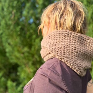 May include: A tan crochet cowl scarf is worn around the neck. The scarf has a textured pattern and is made of a soft, warm material. The person is wearing a dark purple shirt. The background is a blurred green.