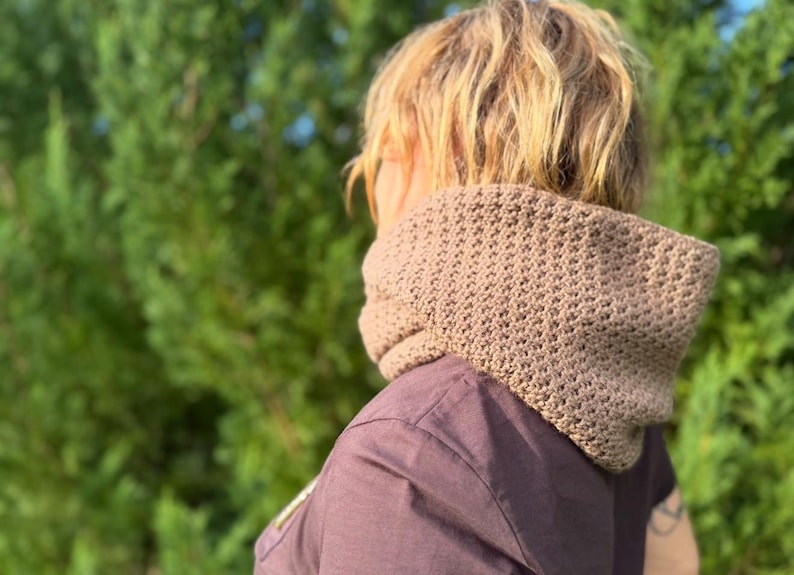 May include: A tan crochet cowl scarf is worn around the neck. The scarf has a textured pattern and is made of a soft, warm material. The person is wearing a dark purple shirt. The background is a blurred green.