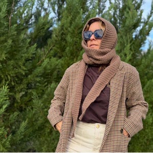 May include: A brown knitted scarf and hood set worn with a brown and tan plaid blazer. The person is wearing sunglasses and a dark brown shirt. The background is a green, leafy tree.