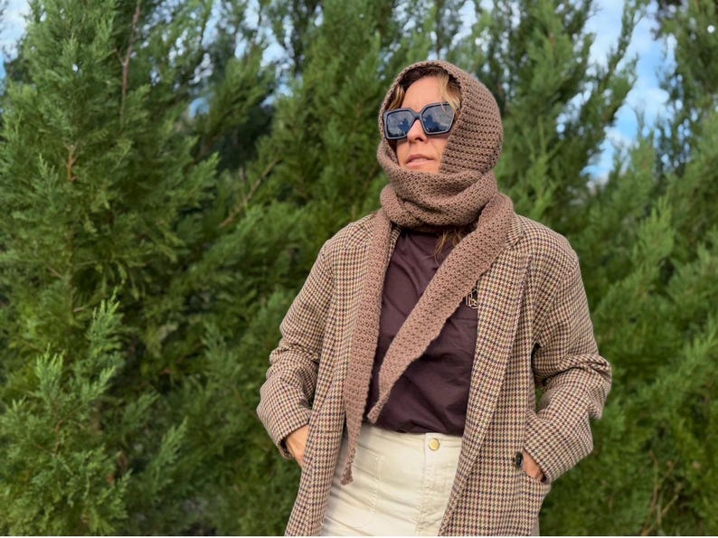 May include: A brown knitted scarf and hood set worn with a brown and tan plaid blazer. The person is wearing sunglasses and a dark brown shirt. The background is a green, leafy tree.