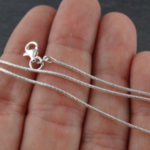 May include: A silver chain necklace with a lobster clasp. The chain is a delicate, fine, and flat snake chain.