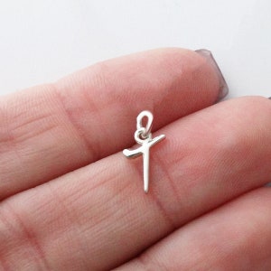 May include: A small, silver-colored cross pendant. The cross has a simple, minimalist design with a loop at the top for a chain. The pendant is held between fingers, showing its size and detail. The background is a soft, neutral color.