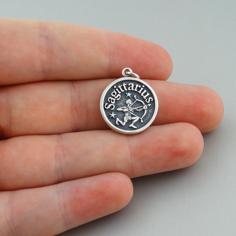 May include: Silver charm with a Sagittarius zodiac symbol. The charm features a round disc with the word "Sagittarius" written around the edge and a depiction of a centaur holding a bow and arrow in the center.