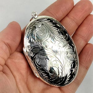 May include: Oval silver locket with intricate floral engraving. The locket has a polished finish and a small loop at the top for a chain. The design features detailed swirling patterns and a dark background.