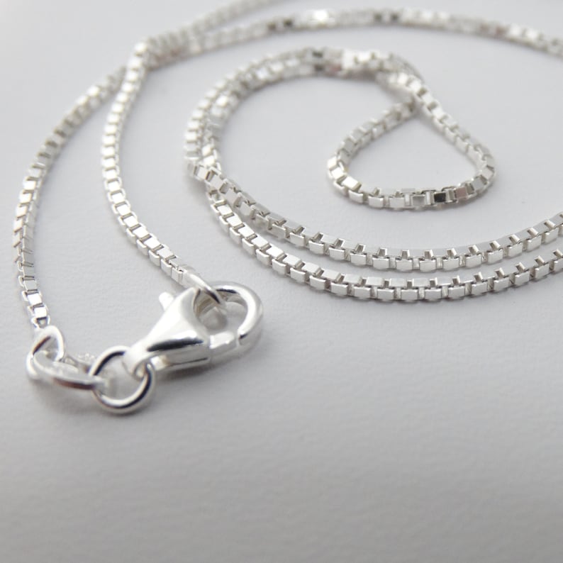May include: A silver chain necklace with a box clasp. The chain is made of small, square links.