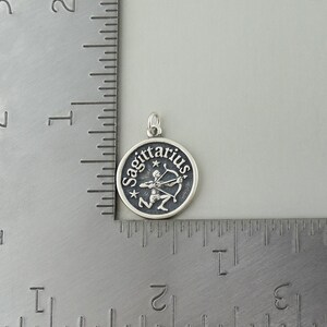 May include: Silver Sagittarius zodiac charm pendant with a black background and a silver archer with a bow and arrow. The charm is about 1 inch in diameter.
