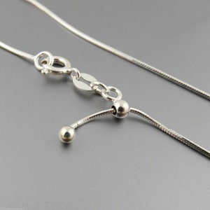 May include: A close-up of a silver snake chain necklace with a lobster clasp and adjustable bead detail. The necklace is displayed on a gray background, showcasing its reflective surface and delicate design. Jewelry.