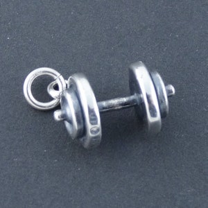 May include: A silver dumbbell charm with a ring for attaching to a necklace or bracelet. The charm is detailed with a textured surface and a small ring at the top.