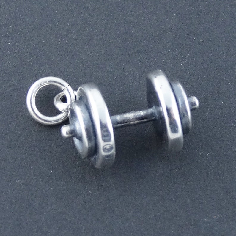 May include: A silver dumbbell charm with a ring for attaching to a necklace or bracelet. The charm is detailed with a textured surface and a small ring at the top.