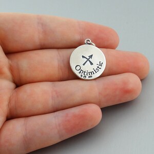 May include: Silver charm with a Sagittarius zodiac symbol and the word "Optimistic" engraved on it.