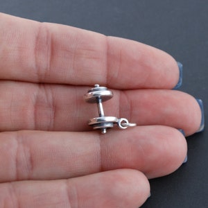 May include: A silver dumbbell charm pendant. The charm is a detailed representation of a dumbbell with a smooth, polished finish.