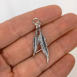 May include: A small, silver-colored feather charm with two detailed feathers. The feathers are connected at the top with a small loop. The charm is designed to be used as a pendant or earring.