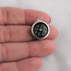 May include: A small silver compass charm with a black face and green markings. The compass face has the cardinal directions N, E, S, and W.