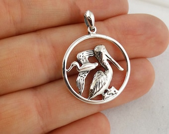 May include: Silver pendant featuring a pelican and a smaller bird inside a circular frame. The pelican is depicted with its beak open, as if feeding its young.