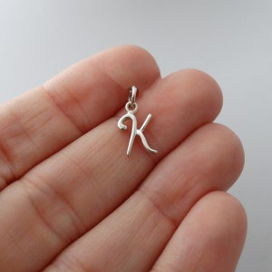 May include: A small, silver-colored pendant in the shape of the letter "K" in a cursive script. The pendant has a loop at the top for attaching to a chain or bracelet. The background is a light, neutral color.