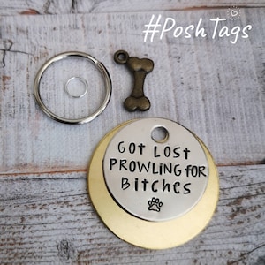Puede incluir: Una placa de identificación para perro de oro y plata con el texto "Got Lost Prowling for Bitches" y una huella de pata. La placa está unida a un llavero plateado con un dije en forma de hueso.