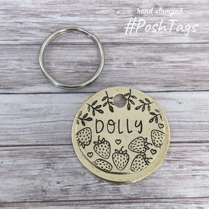 Pode incluir: Uma etiqueta de identificação para cães em metal dourado com um design gravado em preto de morangos, folhas e o nome "DOLLY". A etiqueta está presa a um chaveiro prateado.