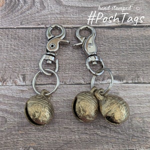 May include: Two metal keychains with silver clasps and split rings. Each keychain has a pair of small, textured, bronze-coloured bells. The words "hand stamped" and "#PoshTags" are printed in the upper right corner.