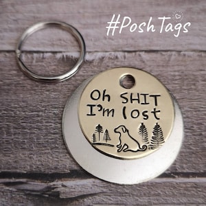 Puede incluir: Una placa de identificación de perro dorada con el texto "Oh Shit I'm Lost" y una silueta de un perro y árboles. La placa está unida a un llavero plateado.