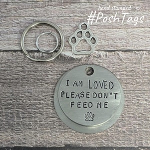 May include: Silver dog tag with the text "I am loved please don't feed me" and a paw print. The tag is attached to a silver key ring with a smaller silver ring.