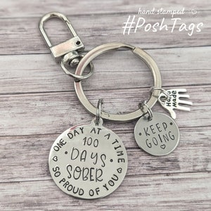 Può includere: Portachiavi d'argento con tre ciondoli. Il ciondolo più grande è rotondo e dice "One day at a time. 100 days sober. So proud of you." Il secondo ciondolo è rotondo e dice "Keep going." Il terzo ciondolo è una piccola mano con le parole "Hand Made".