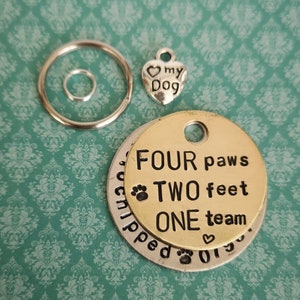 Op de afbeelding: Een gouden en zilveren hondenpenning met de tekst "FOUR paws TWO feet ONE team" en een klein hartvormig sieraad. De penning is bevestigd aan een zilveren sleutelhanger met een kleinere zilveren ring.