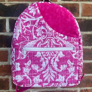 May include: A pink and white floral patterned backpack with a white zipper and a solid pink top section. The backpack has a brown handle and is hanging against a brick wall.