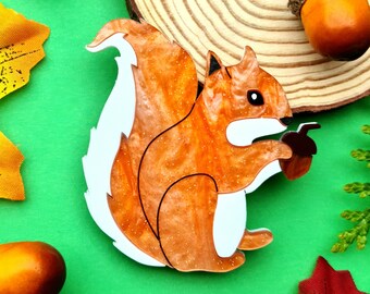 May include: A wooden squirrel-shaped brooch with orange and white details, holding an acorn. The brooch is surrounded by fall-themed decorations, including acorns, leaves, and a wood slice, all set against a green background.