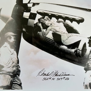 May include: Black and white photograph of a World War II era fighter plane with a pin-up girl painted on the side. Two men stand near the plane, one with his hand on his hip and the other with his hand on his chin. The plane is signed "Charles E.  Shuckeldean 362nd FS 367th FG".