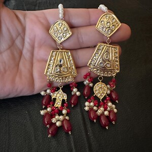 May include: A pair of gold earrings with red and white beads. The earrings have a teardrop shape with intricate designs and are adorned with small, clear stones.