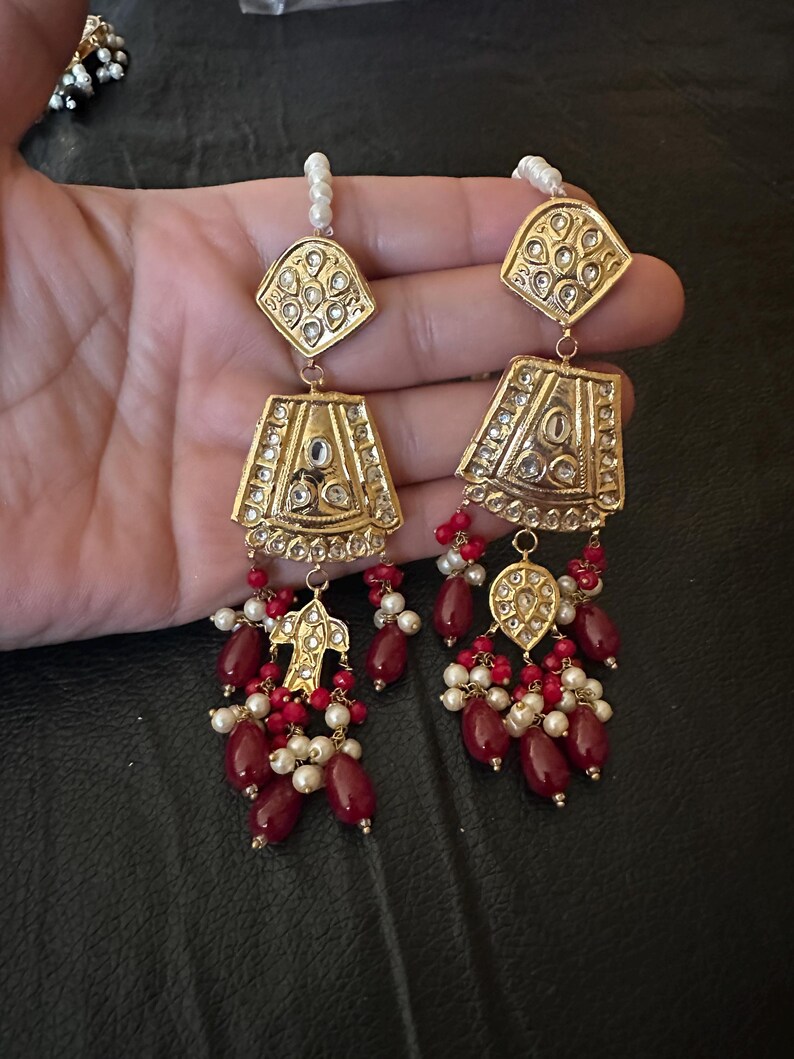 May include: A pair of gold earrings with red and white beads. The earrings have a teardrop shape with intricate designs and are adorned with small, clear stones.