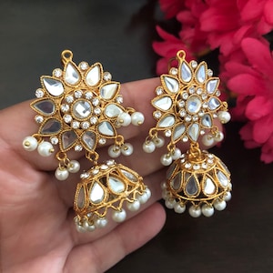 May include: A pair of gold-tone earrings with a mirror-like finish and white pearl accents. The earrings feature a round, floral design with a central stone and a dangling bell-shaped charm.