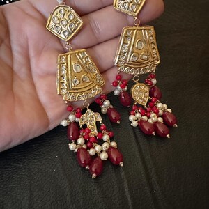 May include: A pair of gold earrings with red and white beads. The earrings have a geometric design with intricate details.
