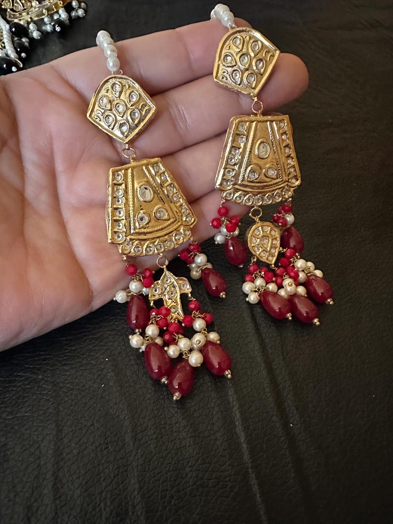 May include: A pair of gold earrings with red and white beads. The earrings have a geometric design with intricate details.