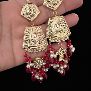 May include: Gold earrings with intricate designs and red and white beads. The earrings feature a geometric shape with a central stone and are adorned with smaller stones.