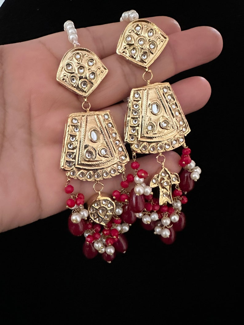 May include: Gold earrings with intricate designs and red and white beads. The earrings feature a geometric shape with a central stone and are adorned with smaller stones.