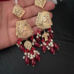 May include: Gold earrings with intricate designs and red and white beads. The earrings feature a teardrop shape with a gold pendant and a dangling chain of beads.