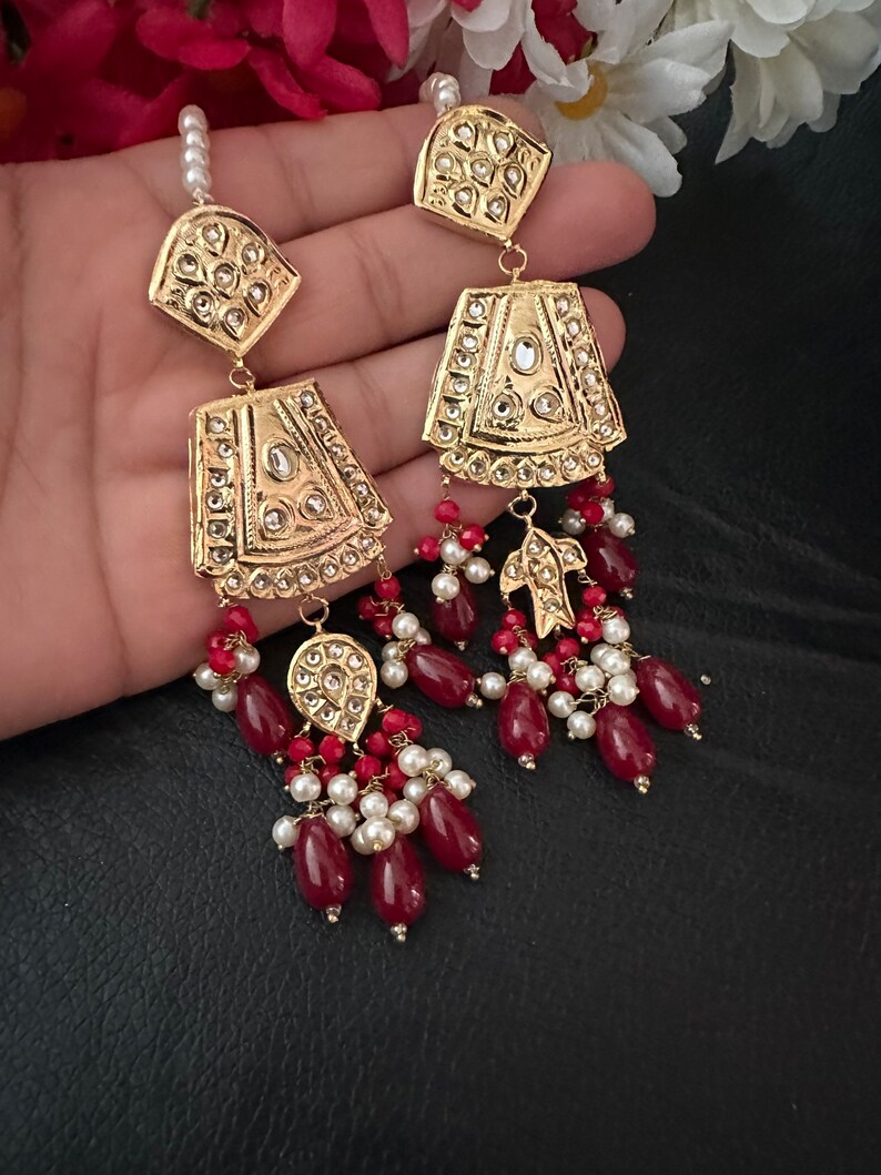 May include: Gold earrings with intricate designs and red and white beads. The earrings feature a teardrop shape with a gold pendant and a dangling chain of beads.