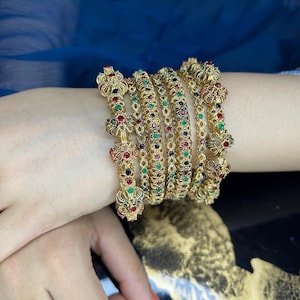May include: A set of five gold-toned metal bangles featuring intricate filigree patterns and embedded small round red, green, and blue stones. The bangles are arranged in a stack, with the outer pieces displaying larger decorative floral-like motifs.