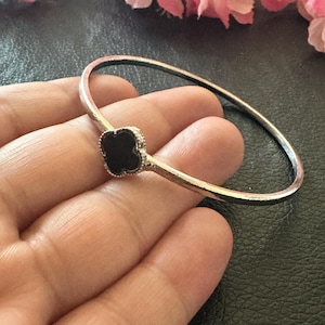 May include: Silver-toned bangle bracelet featuring a black clover-shaped charm. The bracelet is simple and elegant, with a smooth, polished finish. The charm is centered on the bracelet, adding a touch of sophistication. The bracelet is a classic piece of jewelry.