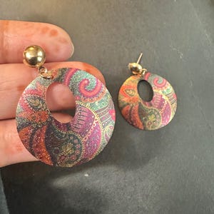 May include: A pair of gold-toned earrings with a circular design. The earrings feature a paisley pattern in shades of red, green, and purple. The earrings have a small gold ball at the top and a large open circle design.