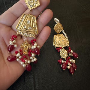 May include: A pair of gold earrings with red and white beads. The earrings feature intricate designs with small, clear stones.