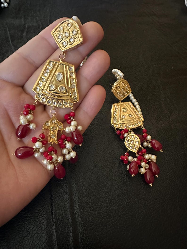 May include: A pair of gold earrings with red and white beads. The earrings feature intricate designs with small, clear stones.